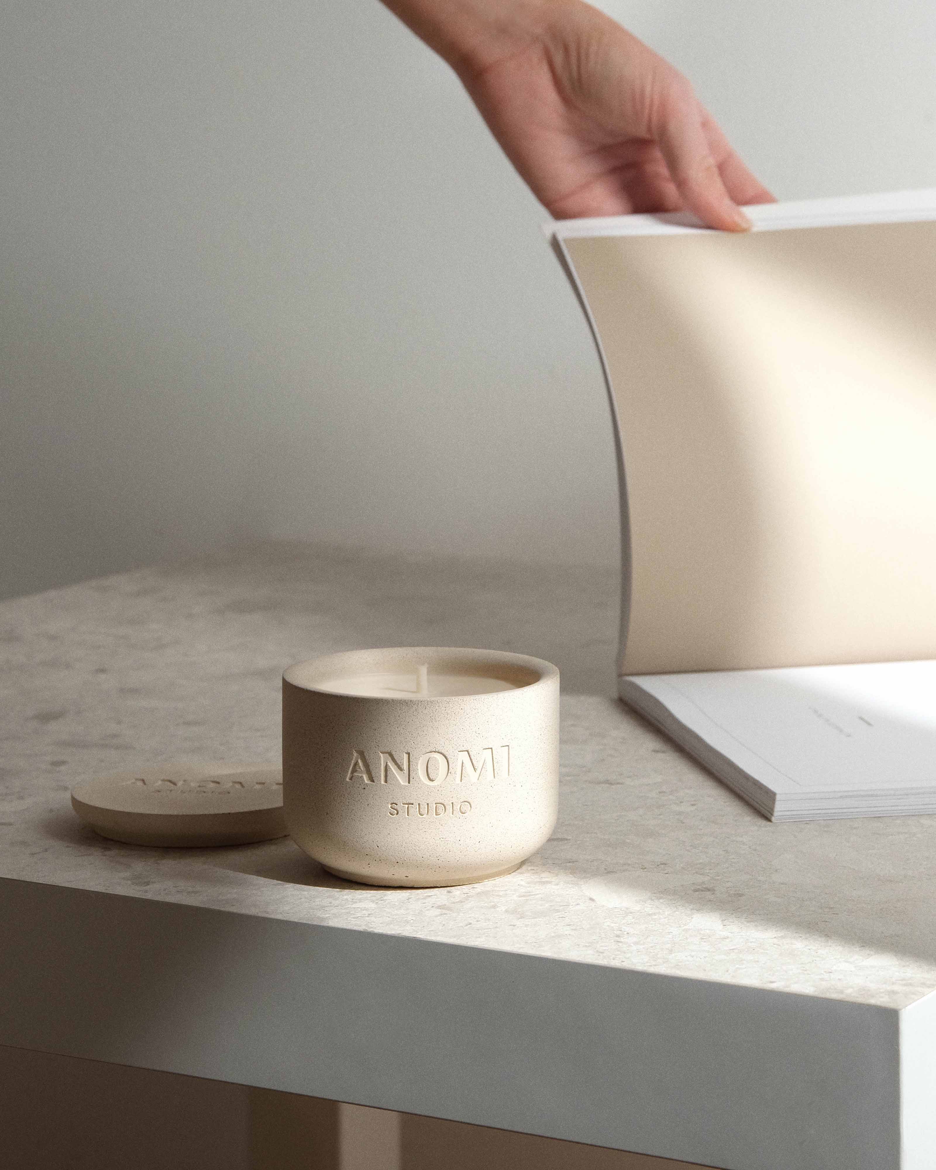 Beige Jesmonite candle with 'ANOMI Studio' branding on a light surface, with a hand and book in the background.